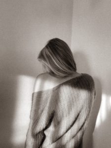 A black and white image of a woman facing a corner, looking down with her sweater falling off her shoulder