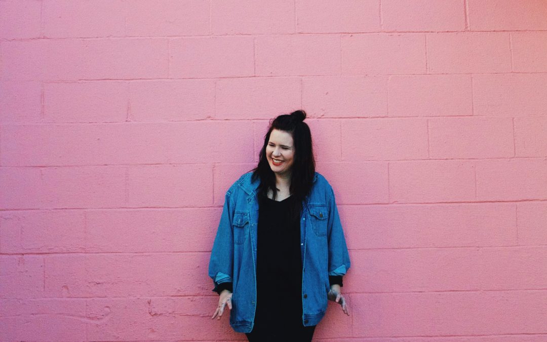 A woman in all black and a jean jacket leans back, laughing against a pink wall