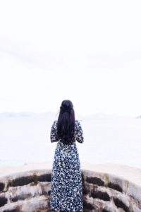 Photo of woman with her back to the camera, in a long dress with long black hair, standing on a stone ledge and looking out into the distance